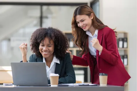 Two women in business attire celebrating success while looking at a laptop. One woman is seated and looking at the screen with her fist raised, while the other stands behind her, placing a hand on her shoulder with her own fist raised.