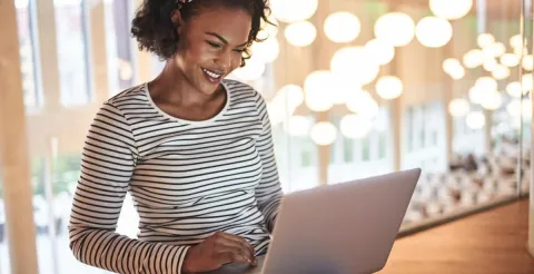 Woman in a striped shirt using a laptop in a brightly lit room, suggesting online research or studying financial aid options.