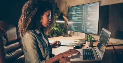 Woman with curly hair works at a desk with a laptop and large monitor displaying code, highlighting female representation in cybersecurity and tech.