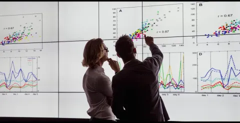 Two people analyze colorful data charts and graphs displayed on a large screen.