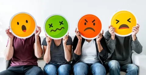Four people sit side by side holding large emoji faces in front of their heads, each emoji showing a different emotion: surprise, sickness, anger, and sadness.