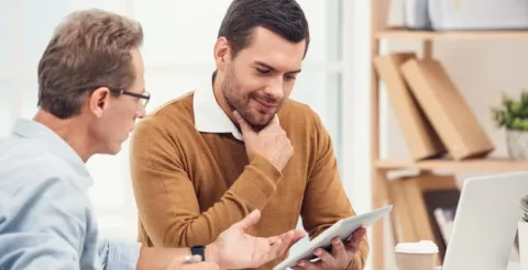 Two men in discussion at a desk, one holding a tablet and the other gesturing, with bookshelves and a laptop in the background, illustrating academic or educational consultation.