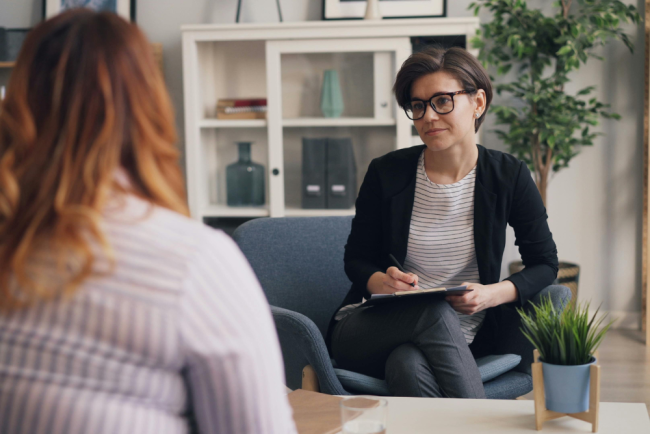 A therapist sits in a chair, holding a clipboard and pen, looking at a patient whose back is to the camera.