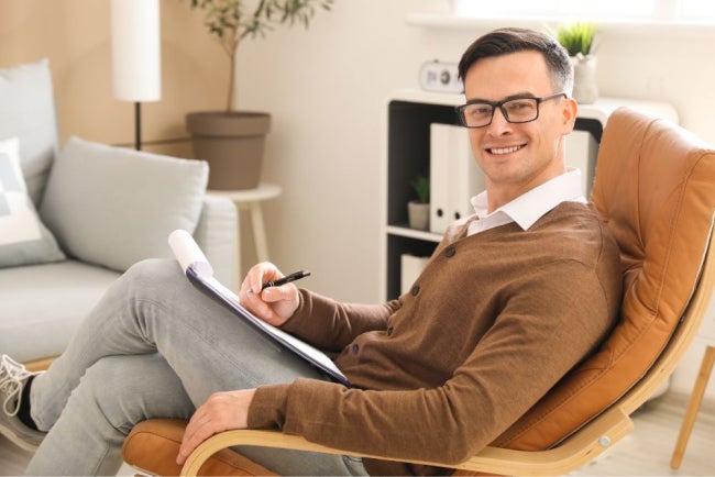 A man wearing glasses and a brown cardigan sits in a leather chair, holding a clipboard and pen.