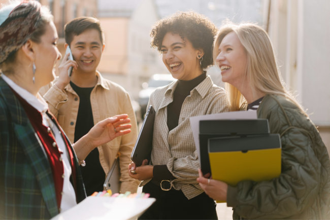 Four people are standing outside and talking. One person is on the phone. Three people are laughing. Two people are holding folders.