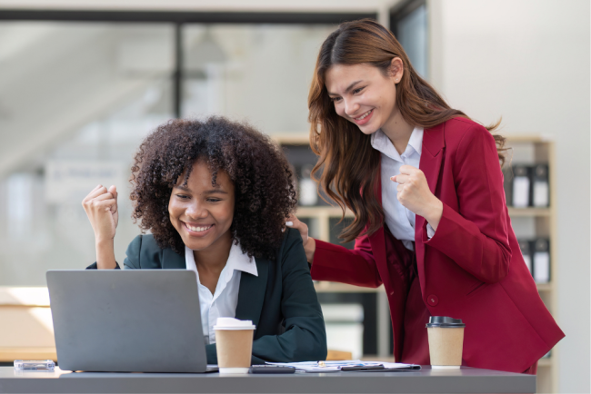 Two women in business attire celebrating success while looking at a laptop. One woman is seated and looking at the screen with her fist raised, while the other stands behind her, placing a hand on her shoulder with her own fist raised.