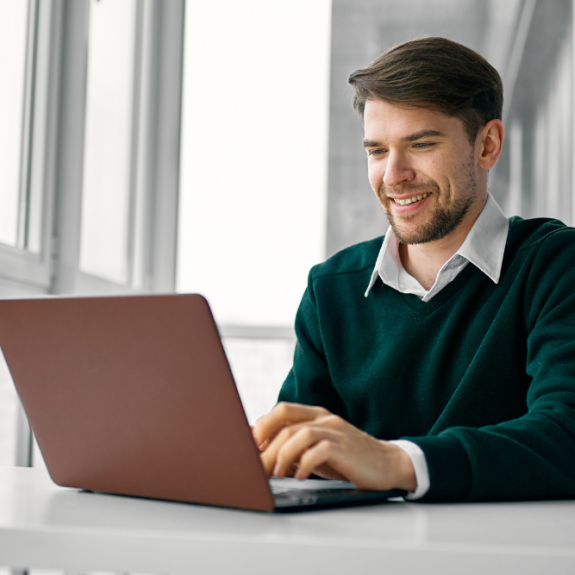 A man wearing a green sweater and white shirt smiles as he types on a maroon laptop at a white desk in front of a window.