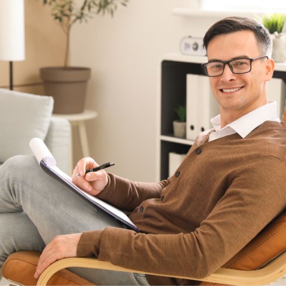 A man wearing glasses and a brown cardigan sits in a leather chair, holding a clipboard and pen.