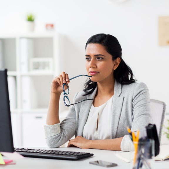 A person sits at a desk with a computer, holding glasses and looking off to the side thoughtfully.