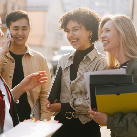 Four people are standing outside and talking. One person is on the phone. Three people are laughing. Two people are holding folders.
