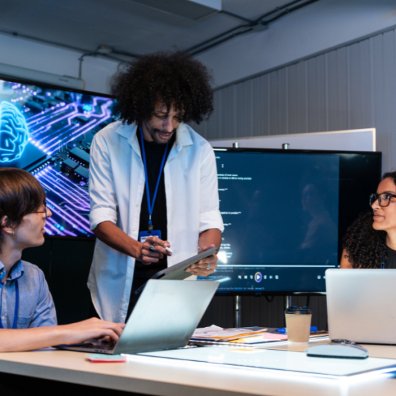 Three people are gathered around a table in a dimly lit room, with a large screen displaying a blue, circuit-board-like brain graphic behind them. 