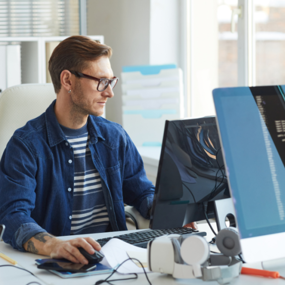 A person wearing glasses and a blue shirt sits at a desk, working on a computer with multiple monitors displaying code.