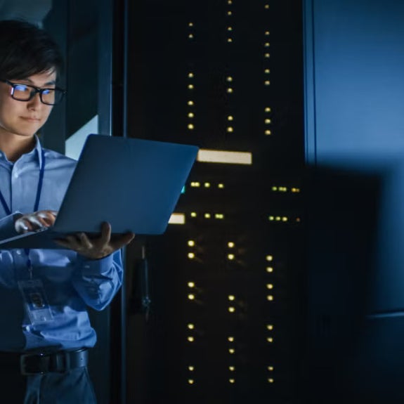 IT professional checking a laptop in a data center with blue lighting and server towers.