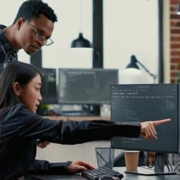 Two people looking intently at a computer screen in an office setting, the woman sitting and pointing while man stands looking at the same screen.