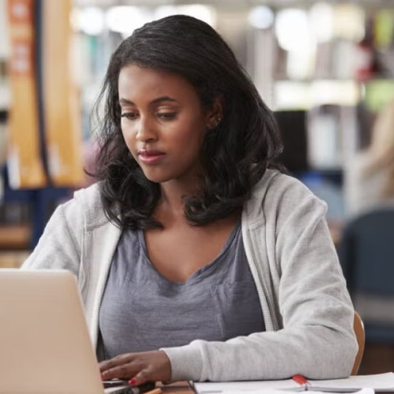 Woman working on a laptop in a public library, focused on her studies, suggesting career advancement or preparation for a finance manager role.