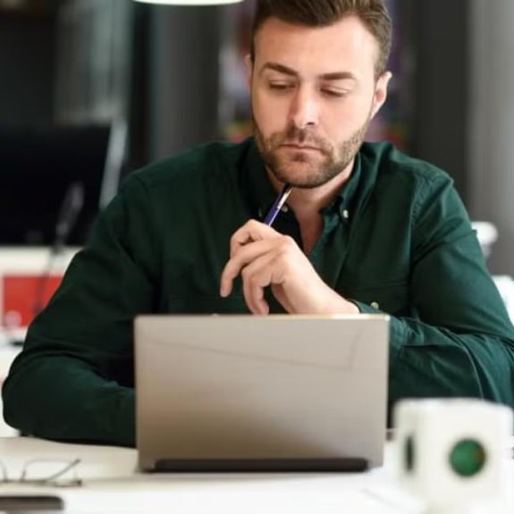 Man in a green shirt sits in a modern office at a desk with a laptop, holding a pen thoughtfully, suggesting focus on analytical work or career planning.