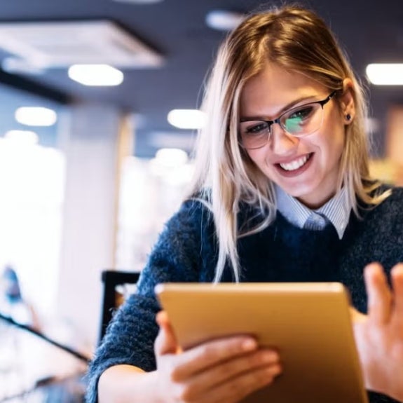 Woman with long blonde hair uses a tablet in a modern cafe or workspace, illustrating participation and advancement of women in the finance sector.
