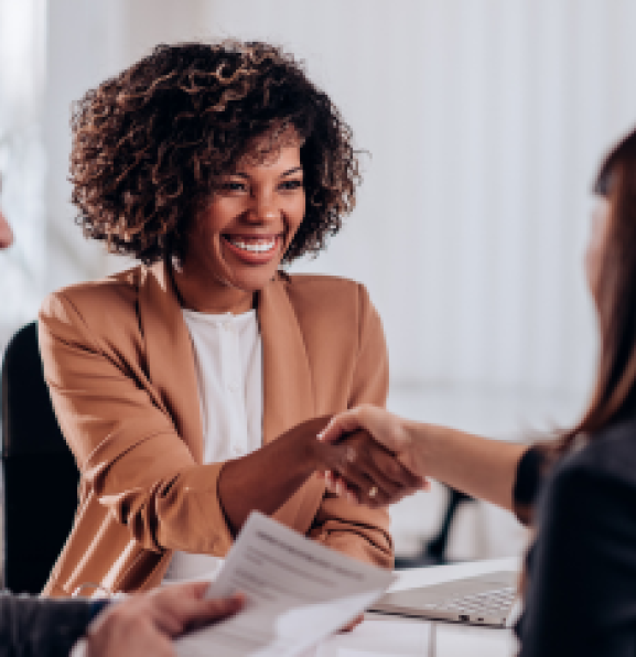 Two women shaking hands in a business meeting while male colleague smiles on the left.