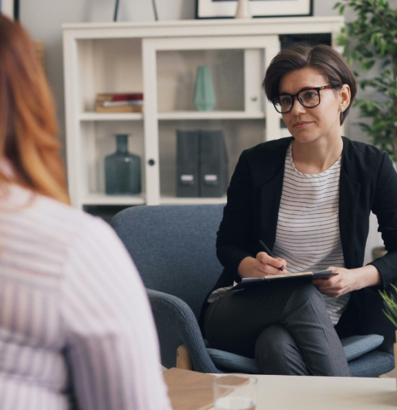 A therapist sits in a chair, holding a clipboard and pen, looking at a patient whose back is to the camera.