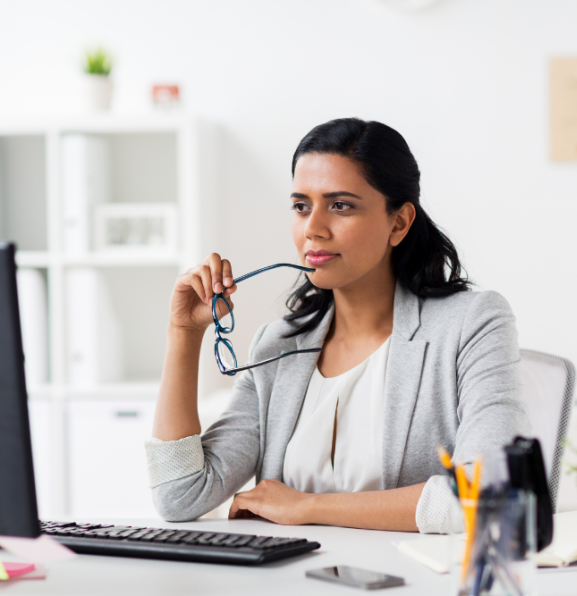A person sits at a desk with a computer, holding glasses and looking off to the side thoughtfully.