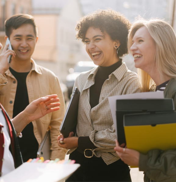 Four people are standing outside and talking. One person is on the phone. Three people are laughing. Two people are holding folders.