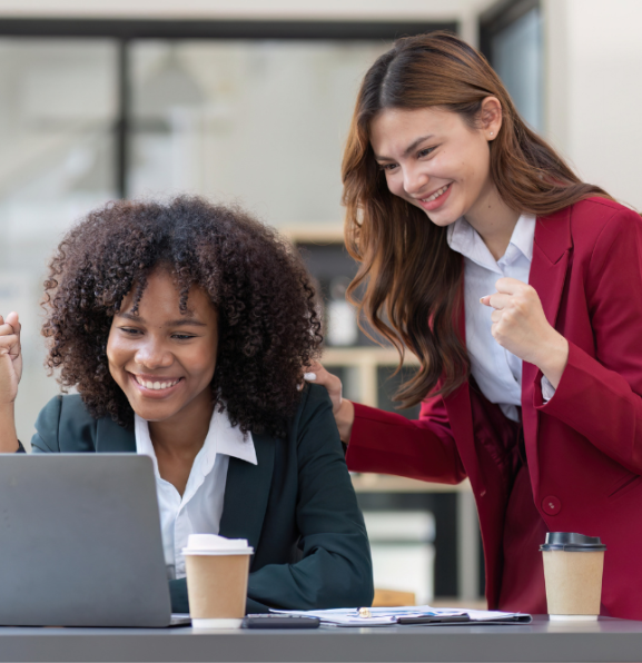 Two women in business attire celebrating success while looking at a laptop. One woman is seated and looking at the screen with her fist raised, while the other stands behind her, placing a hand on her shoulder with her own fist raised.
