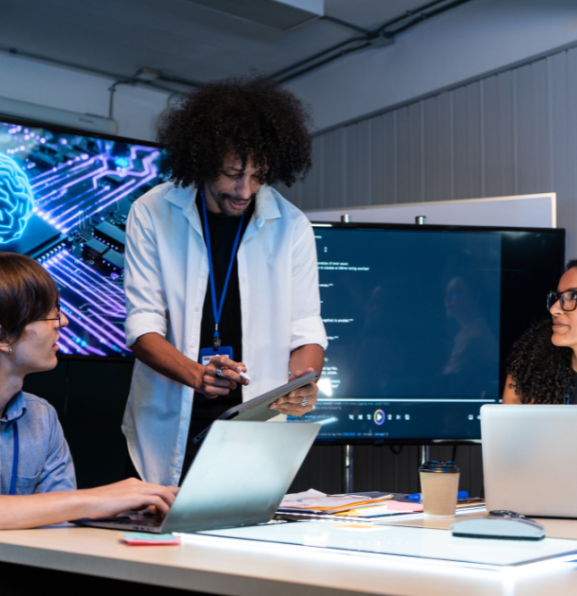 Three people are gathered around a table in a dimly lit room, with a large screen displaying a blue, circuit-board-like brain graphic behind them. 