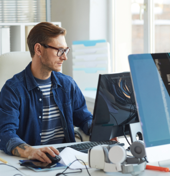 A person wearing glasses and a blue shirt sits at a desk, working on a computer with multiple monitors displaying code.
