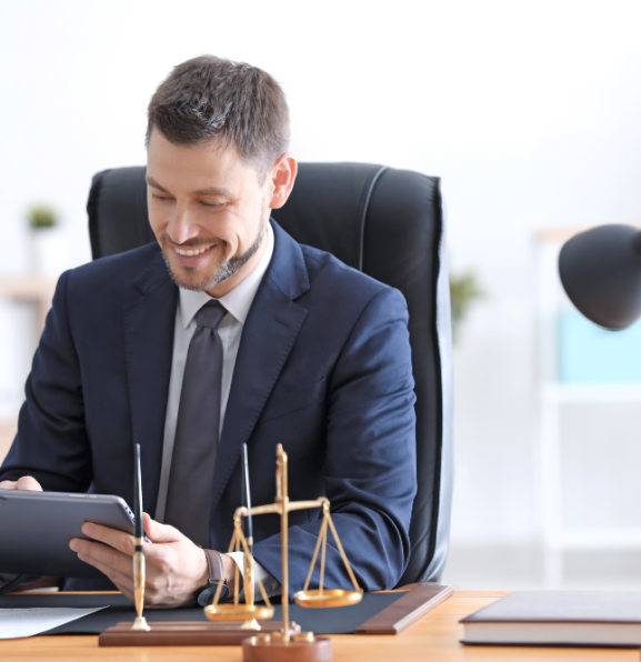 A man in a suit sits at a desk, smiling and looking at a tablet. On the desk are a pen, a scale of justice, and a lamp.