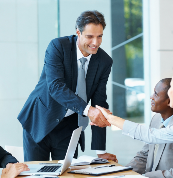 Business meeting in a bright office: a man in a suit stands and smiles while shaking hands with colleagues around a conference table with laptops and documents.