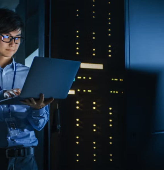 IT professional checking a laptop in a data center with blue lighting and server towers.