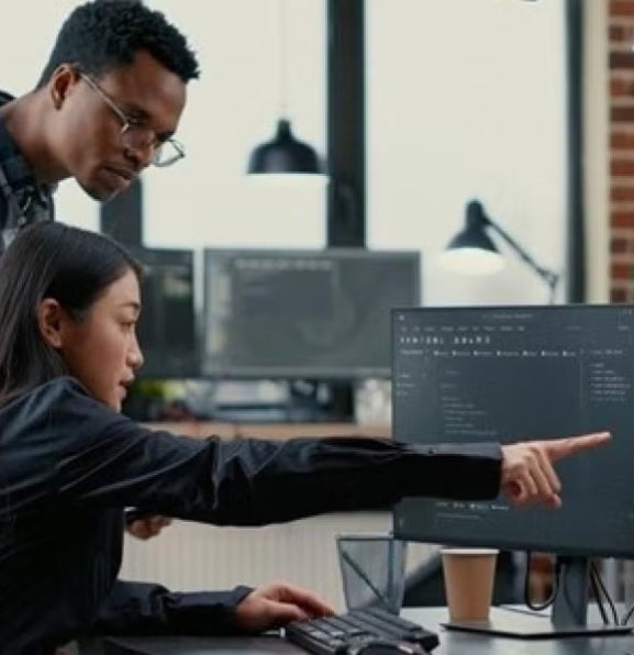 Two people looking intently at a computer screen in an office setting, the woman sitting and pointing while man stands looking at the same screen.