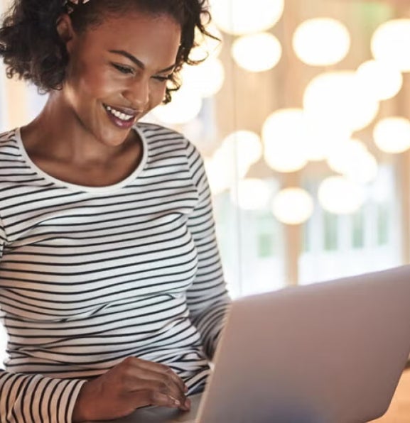 Woman in a striped shirt using a laptop in a brightly lit room, suggesting online research or studying financial aid options.