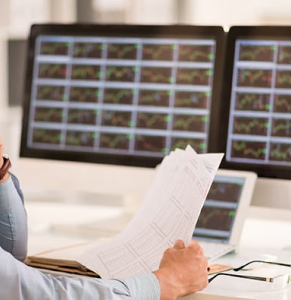 Man in business attire analyzes market data across multiple monitors, holding data sheets, illustrating the rise of marketing analytics in finance.