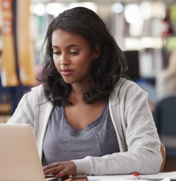 Woman working on a laptop in a public library, focused on her studies, suggesting career advancement or preparation for a finance manager role.