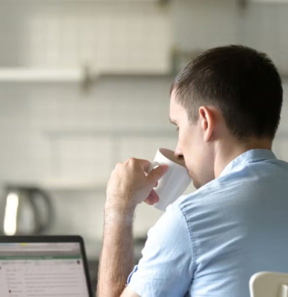 Man in a light blue shirt sits at a kitchen table with a laptop, drinking from a cup, suggesting remote learning or online study in a home environment.