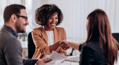 Two women shaking hands in a business meeting while male colleague smiles on the left.