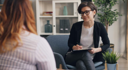 A therapist sits in a chair, holding a clipboard and pen, looking at a patient whose back is to the camera.