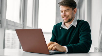 A man wearing a green sweater and white shirt smiles as he types on a maroon laptop at a white desk in front of a window.
