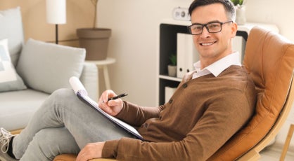 A man wearing glasses and a brown cardigan sits in a leather chair, holding a clipboard and pen.