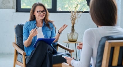 A woman in a denim shirt and glasses holds a clipboard and pen while speaking to another person in a therapy session.