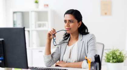 A person sits at a desk with a computer, holding glasses and looking off to the side thoughtfully.