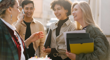 Four people are standing outside and talking. One person is on the phone. Three people are laughing. Two people are holding folders.