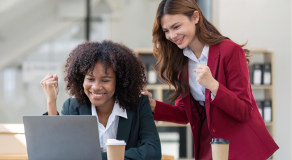 Two women in business attire celebrating success while looking at a laptop. One woman is seated and looking at the screen with her fist raised, while the other stands behind her, placing a hand on her shoulder with her own fist raised.