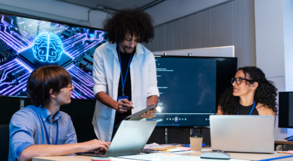 Three people are gathered around a table in a dimly lit room, with a large screen displaying a blue, circuit-board-like brain graphic behind them. 
