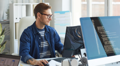 A person wearing glasses and a blue shirt sits at a desk, working on a computer with multiple monitors displaying code.