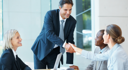 Business meeting in a bright office: a man in a suit stands and smiles while shaking hands with colleagues around a conference table with laptops and documents.