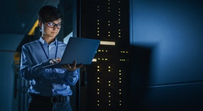 IT professional checking a laptop in a data center with blue lighting and server towers.