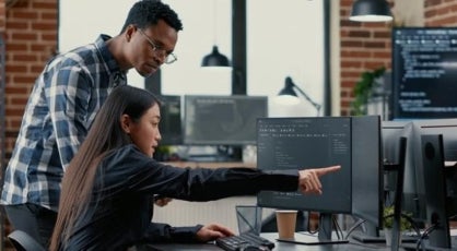 Two people looking intently at a computer screen in an office setting, the woman sitting and pointing while man stands looking at the same screen.