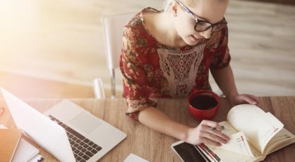 Woman in a floral dress sits at a desk with a planner and coffee, using a laptop, suggesting balancing work tasks and study in a home or casual workspace